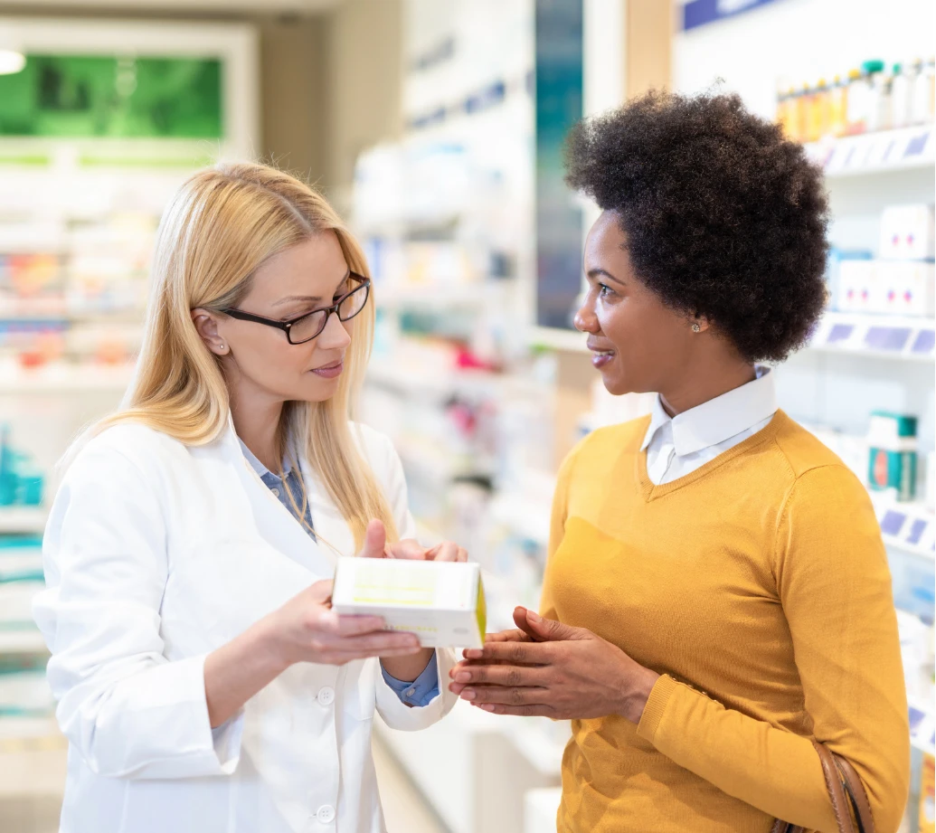 A pharmacist shows a medication box to a woman in a pharmacy, both engaged in conversation.