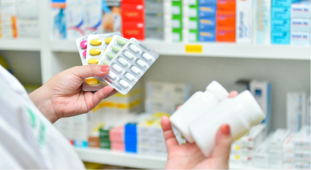 Person holds pill blister packs and medicine bottles in a pharmacy with shelves of medications in the background.
