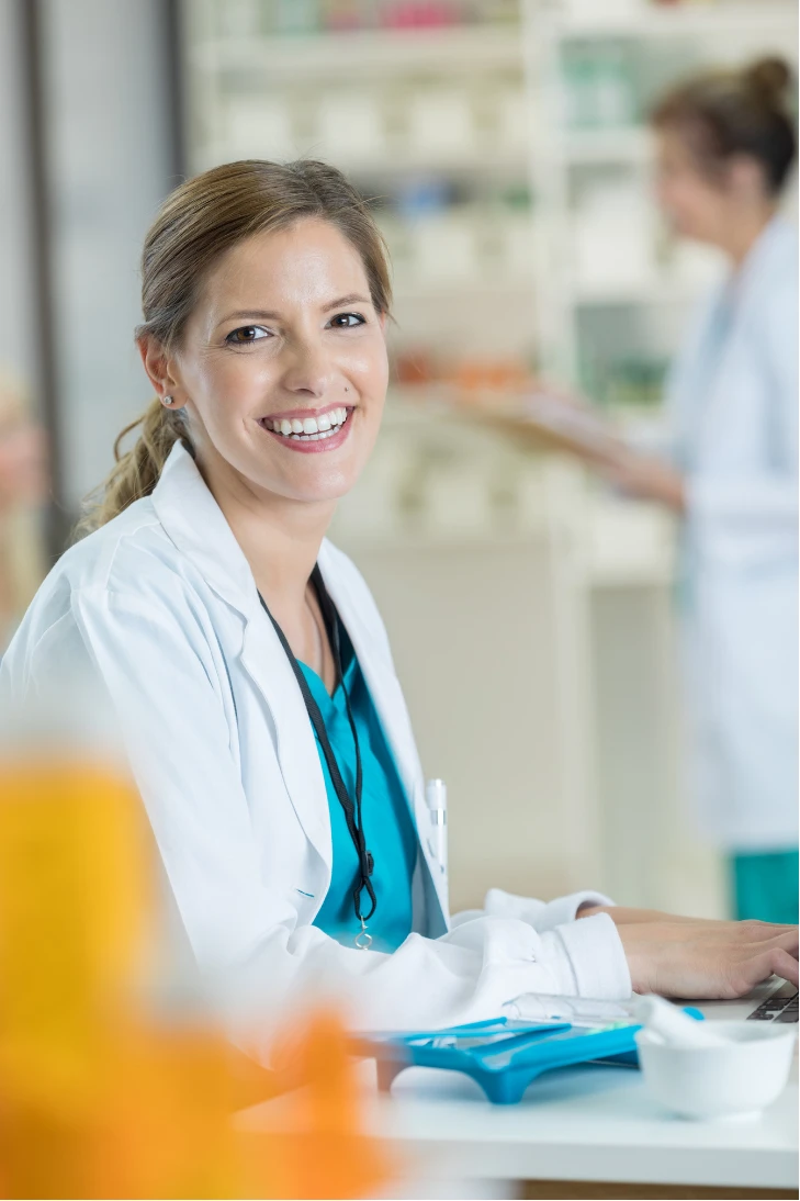 Smiling female pharmacist in a white coat working at a pharmacy counter with shelves in the background.