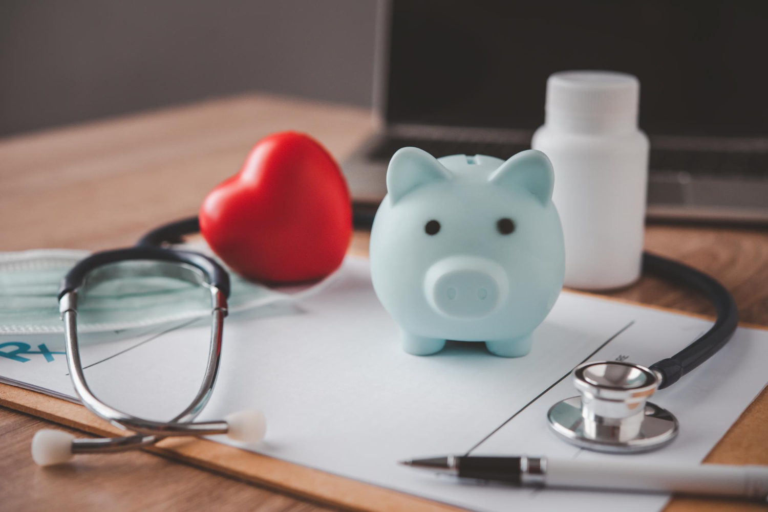 A blue piggy bank, red heart, stethoscope, mask, and medicine bottle on a clipboard and desk.
