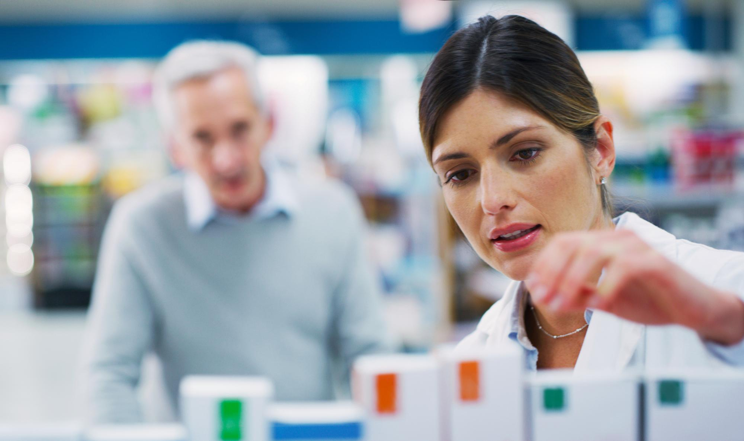 A pharmacist selects medication while an older man waits in the background at a pharmacy counter.