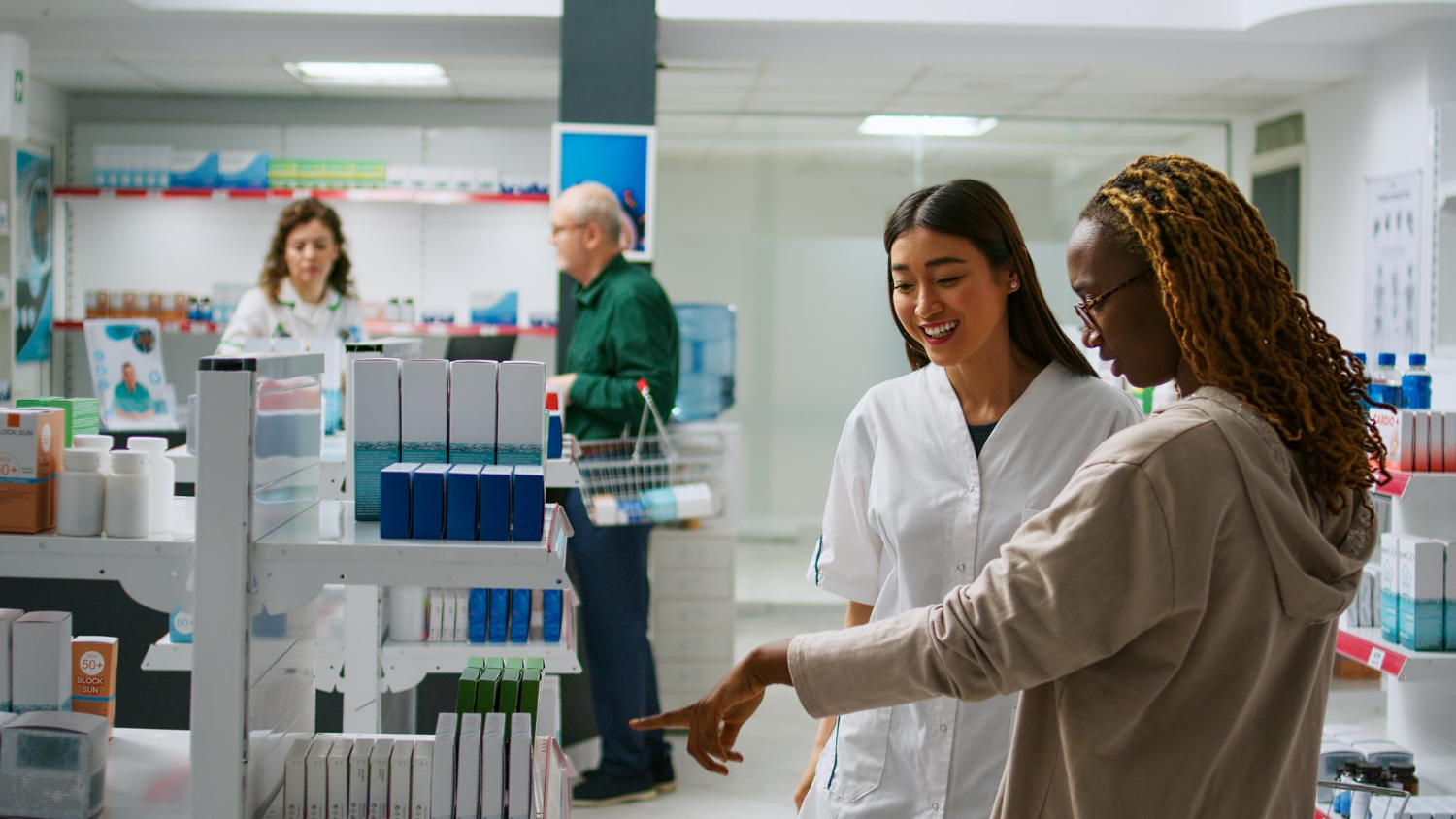 Two women talk while looking at products in a bright pharmacy with shelves and customers in the background.