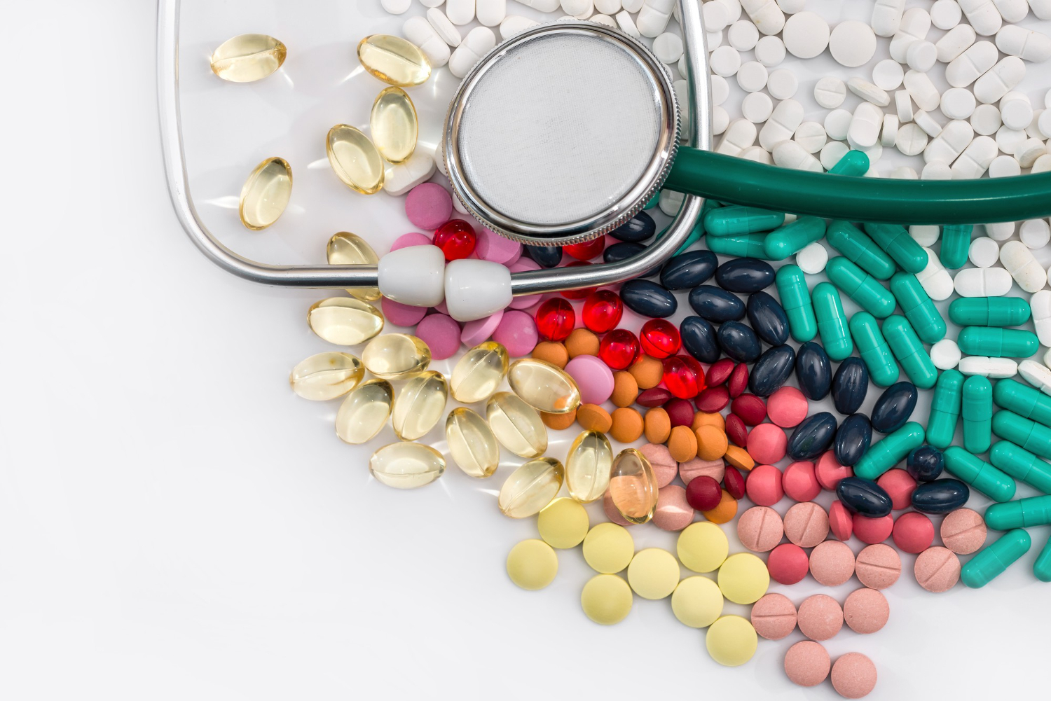 Assorted pills and capsules spread next to a stethoscope on a white background.