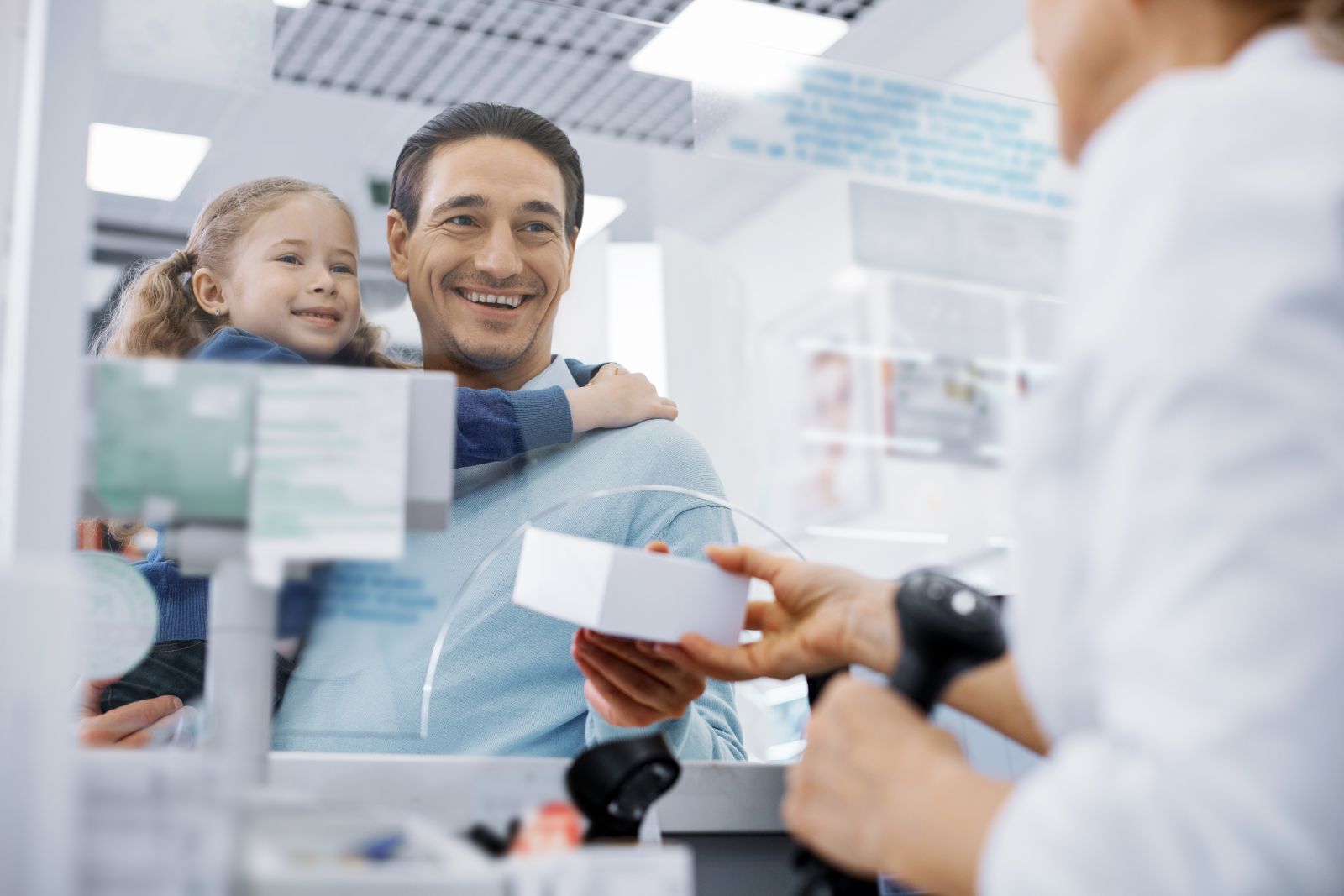 A man holding a smiling child receives a box from a pharmacist at a pharmacy counter.
