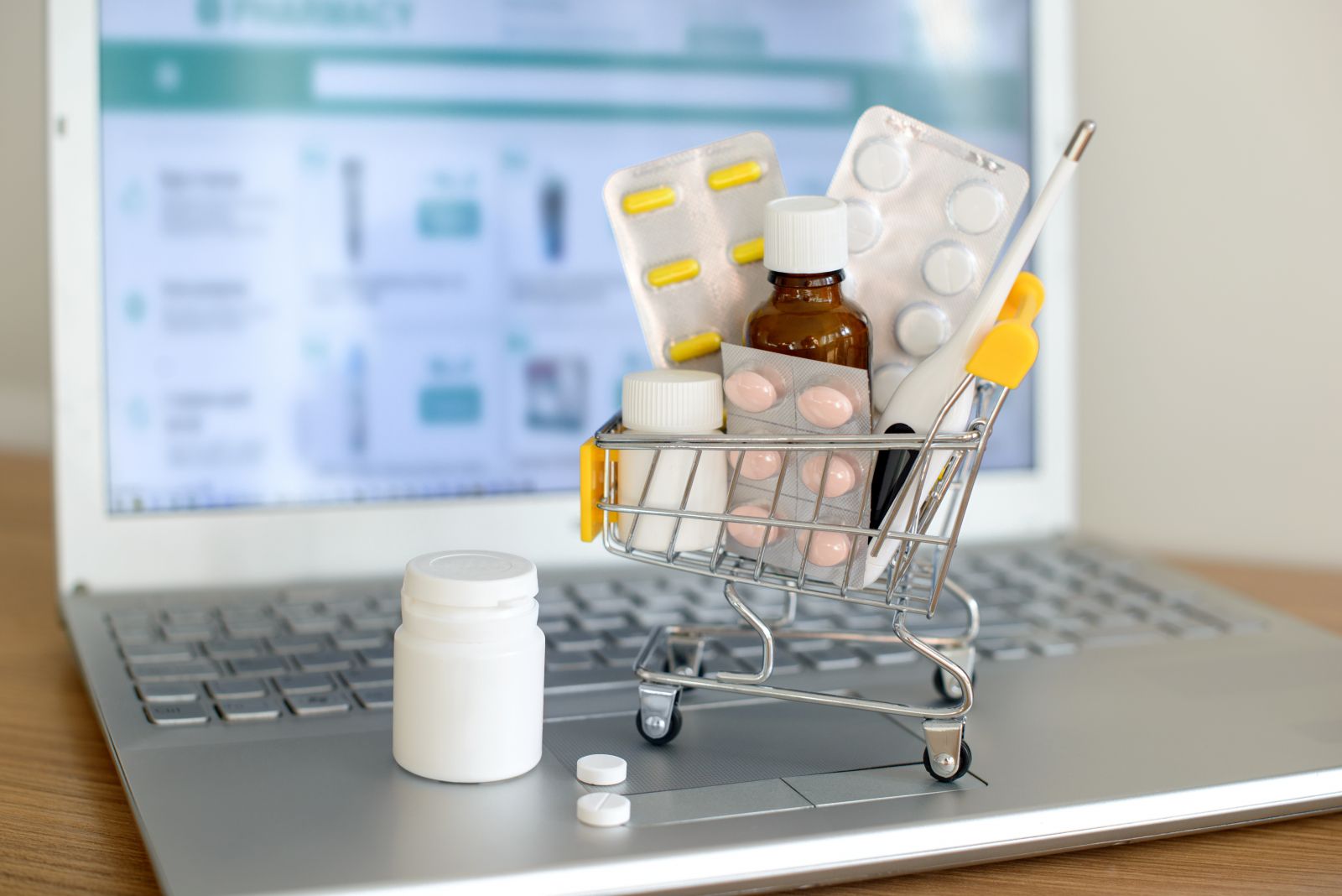 Mini shopping cart with medicine, pills, and thermometer on laptop, symbolizing online pharmacy or medicine shopping.