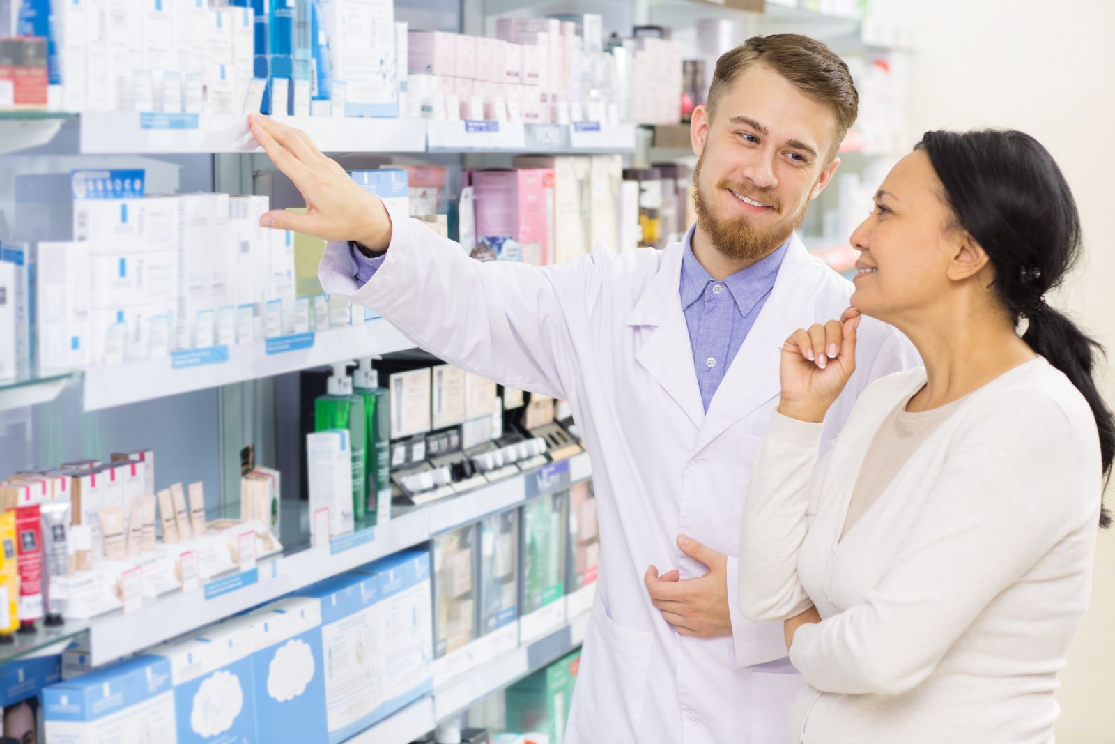 Pharmacist helping a woman choose products at a pharmacy, both smiling and talking by the shelves.