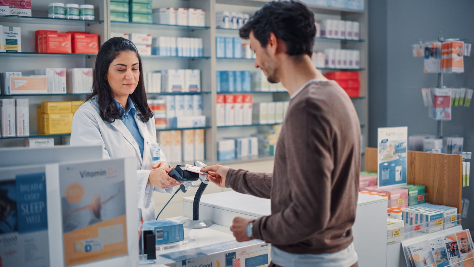 A pharmacist helps a customer paying by card at the pharmacy counter, surrounded by medicines and products.