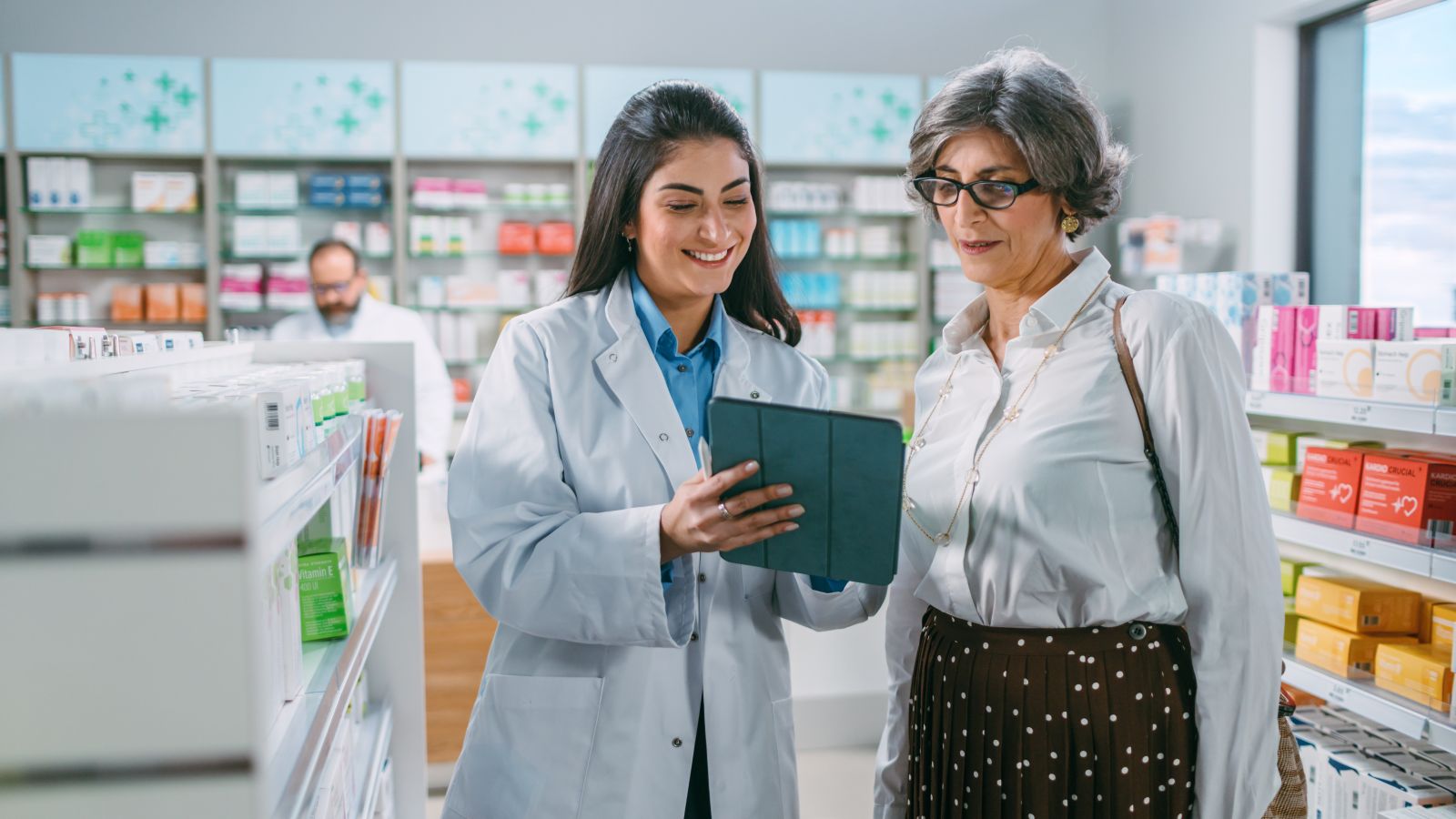 A pharmacist shows a tablet to a customer in a pharmacy aisle, both smiling and engaged in conversation.