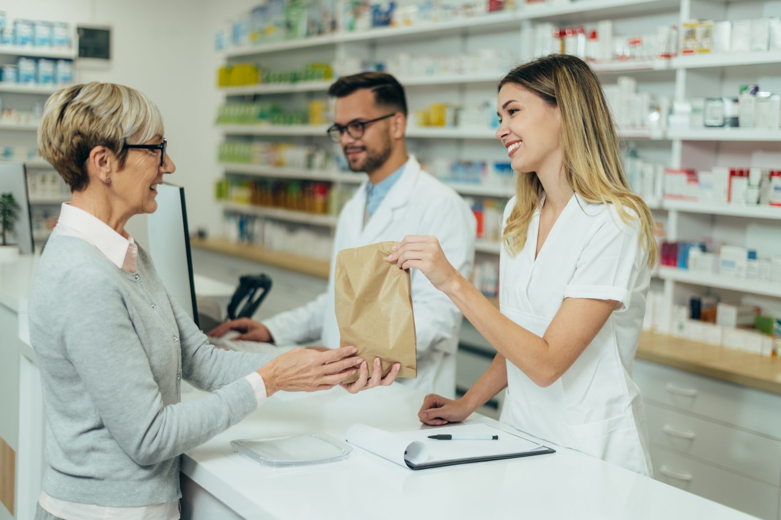 A pharmacist hands a paper bag to a smiling customer at a pharmacy counter, with another pharmacist nearby.