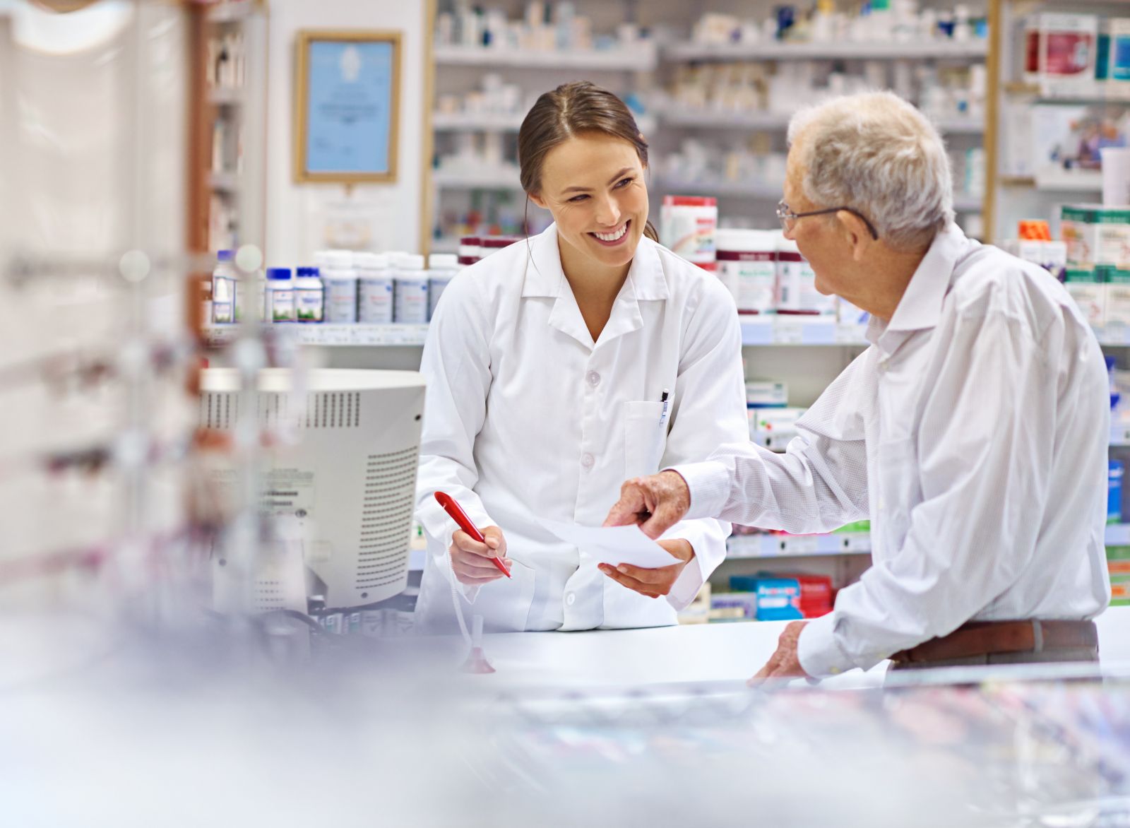 A pharmacist smiling and helping an elderly man with his prescription at a pharmacy counter.