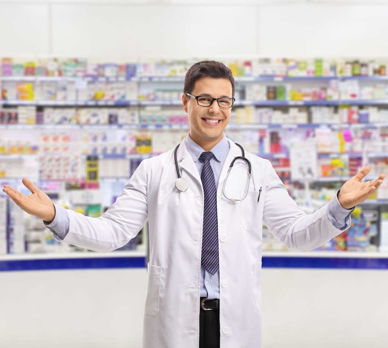 Smiling male doctor with open arms stands in front of shelves filled with pharmaceutical products.