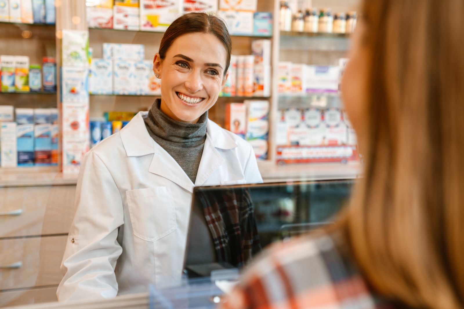 Smiling pharmacist in a white coat helps a customer at the pharmacy counter.