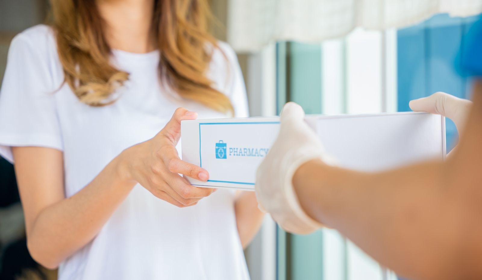 A woman receives a pharmacy package from a gloved delivery person at her door.