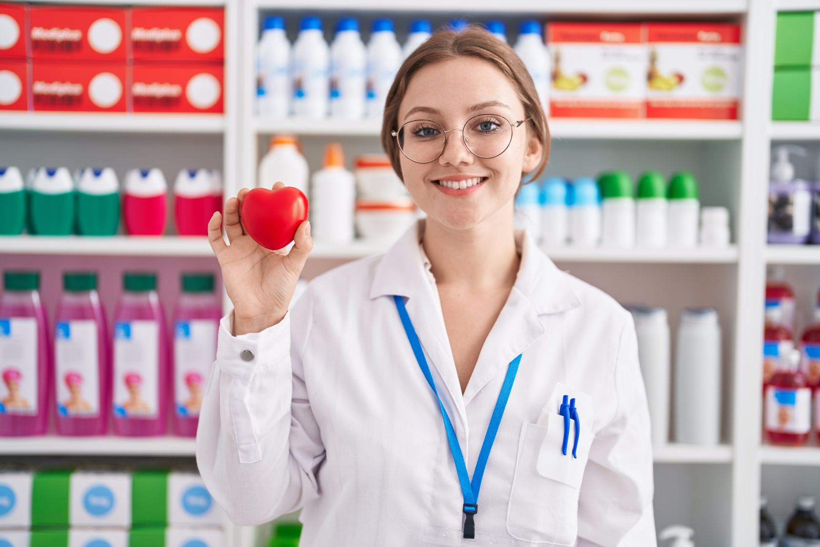 Smiling pharmacist holds a red heart-shaped object in a pharmacy with products on shelves behind her.