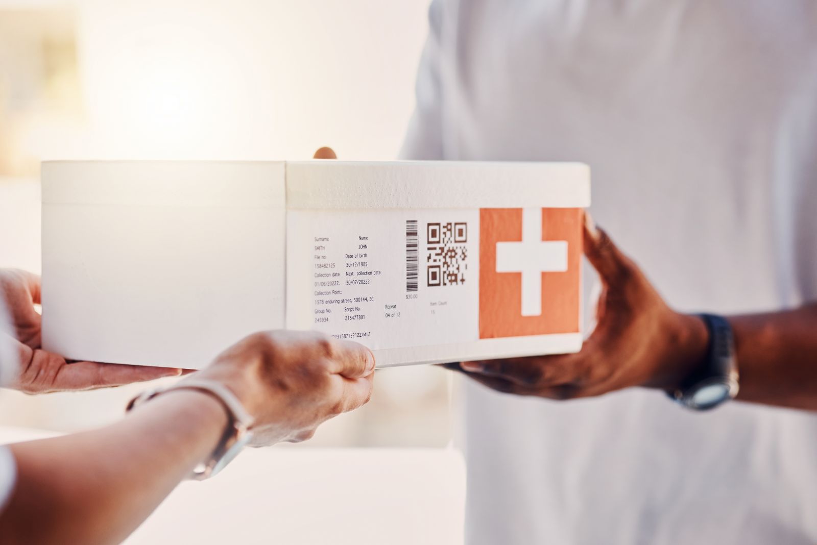 Two people exchange a white medical package with a red label and a white cross.