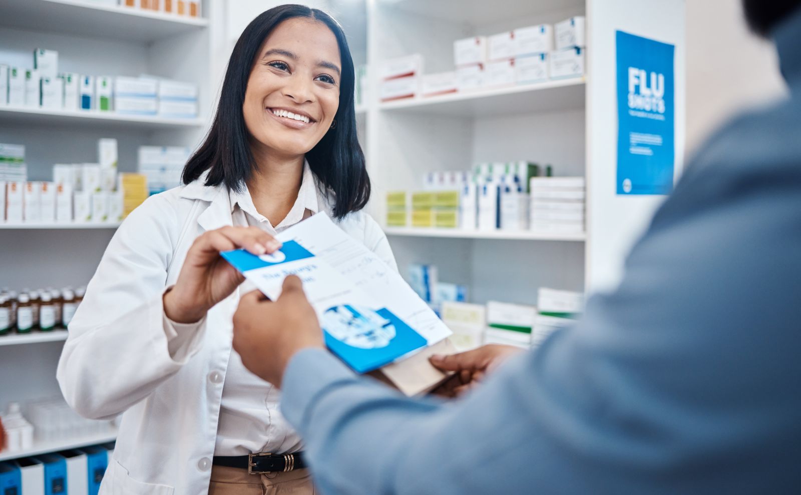 AdobeStock_580025431 Smiling pharmacist hands a prescription bag to a customer at a pharmacy counter.
