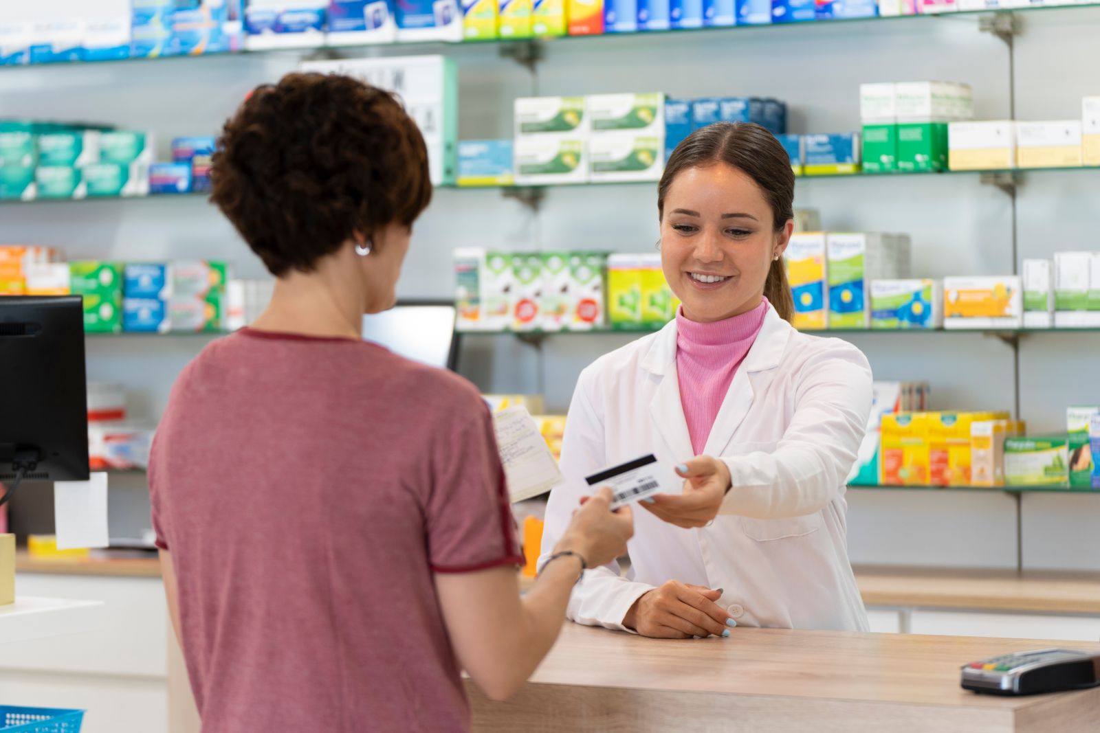 A pharmacist hands a payment card to a customer at a pharmacy counter, with shelves of products behind them.