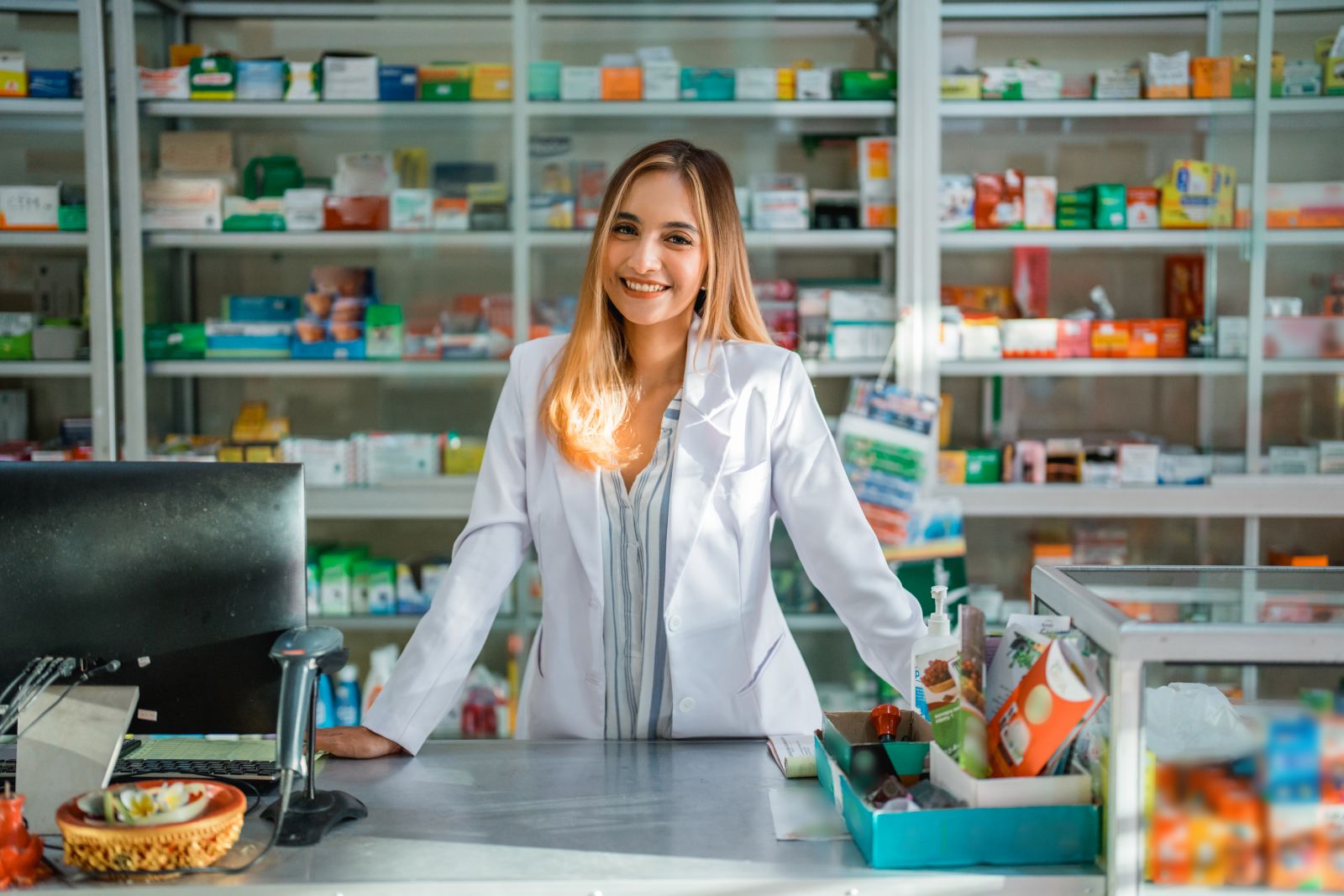Smiling pharmacist stands behind the counter in a pharmacy, surrounded by shelves of medicines.