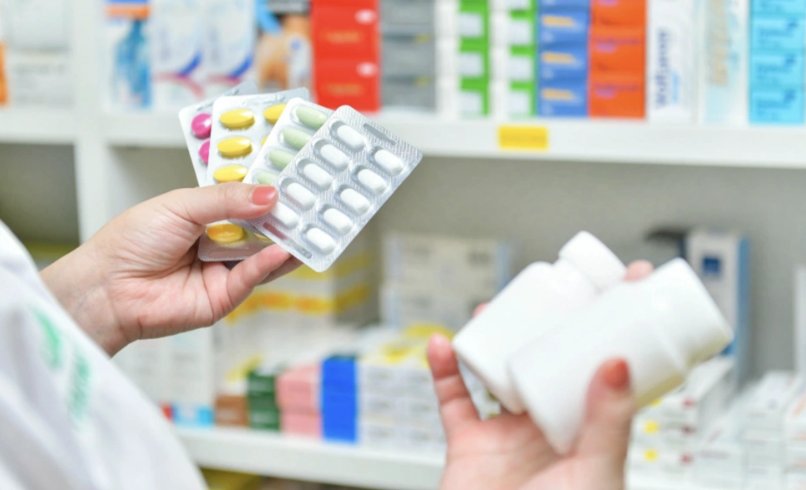 A person holds pill blister packs and medicine bottles in a pharmacy with shelves of medications behind.