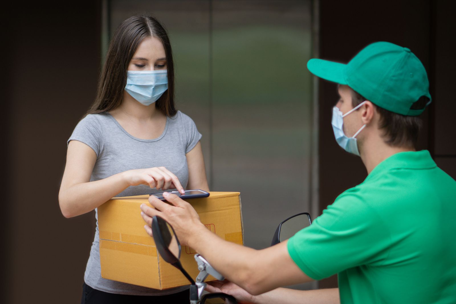 A woman in a mask signs for a package on a delivery person’s device; both are wearing masks.
