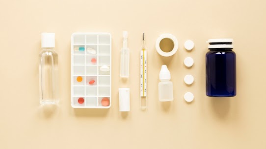 Medical supplies, pills, pill organizer, thermometer, bottle, and tape arranged neatly on a beige surface.