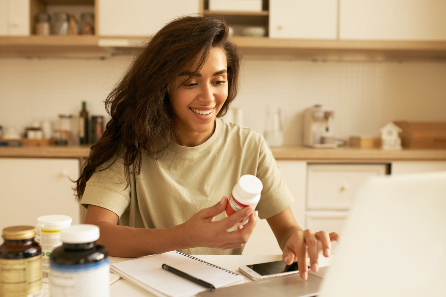 Smiling woman at kitchen table looks at a supplement bottle while working on a laptop with notebook nearby.