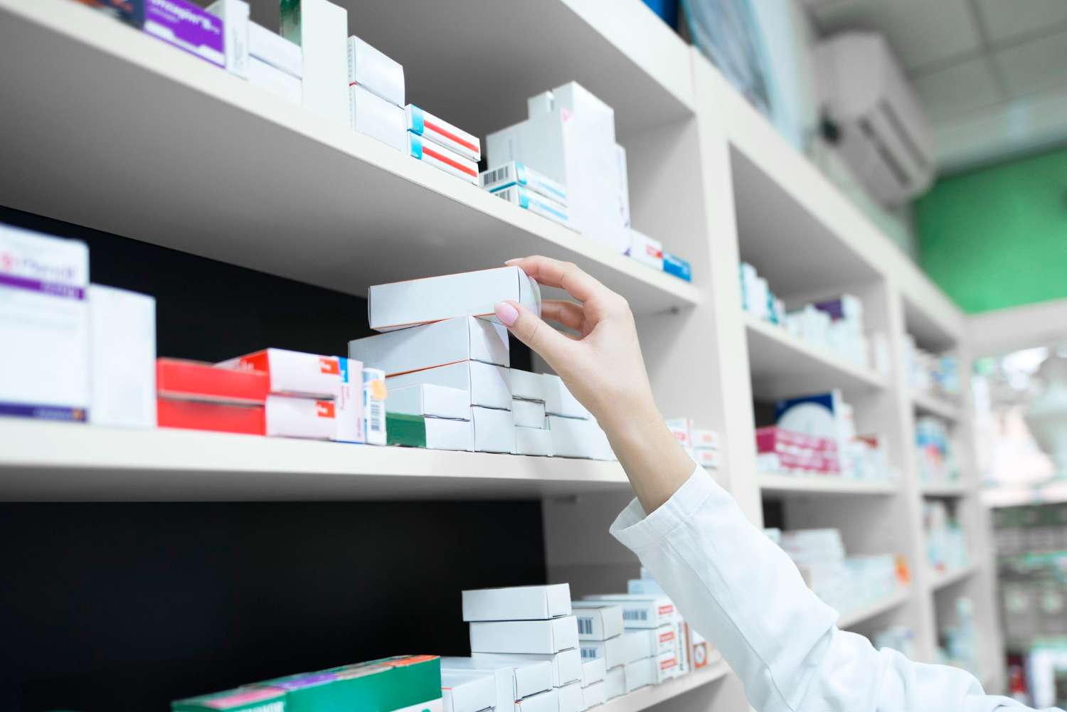 A pharmacist’s hand reaching for a box of medicine on a pharmacy shelf filled with various medications.