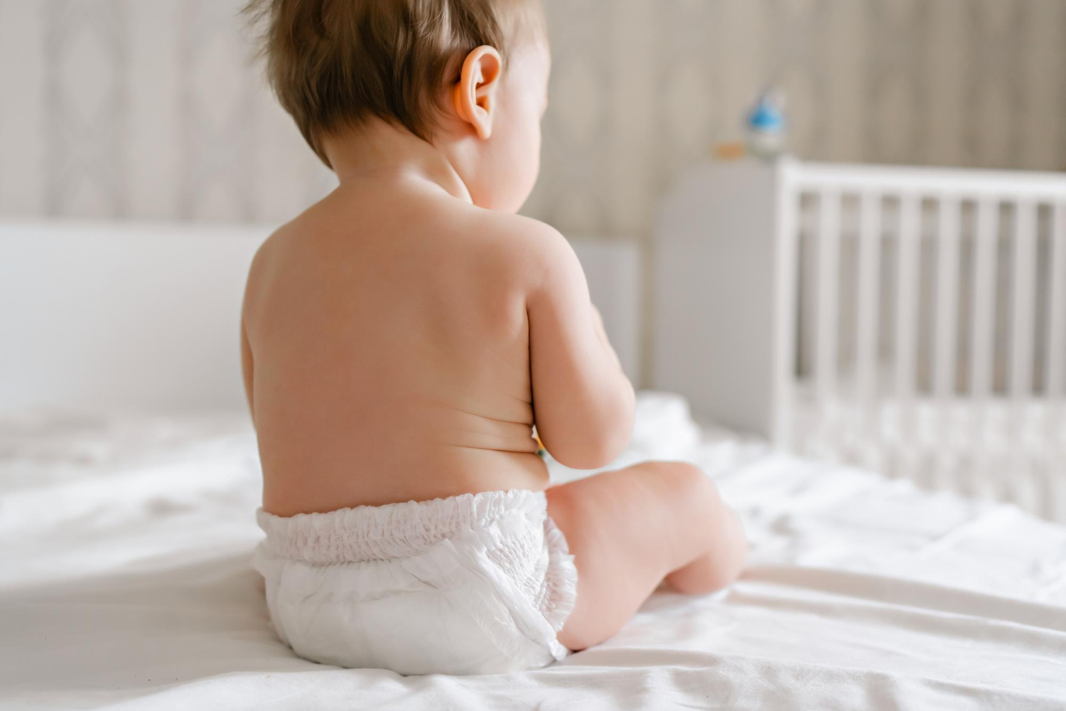 A baby wearing a diaper sits on a bed with a crib in the background.