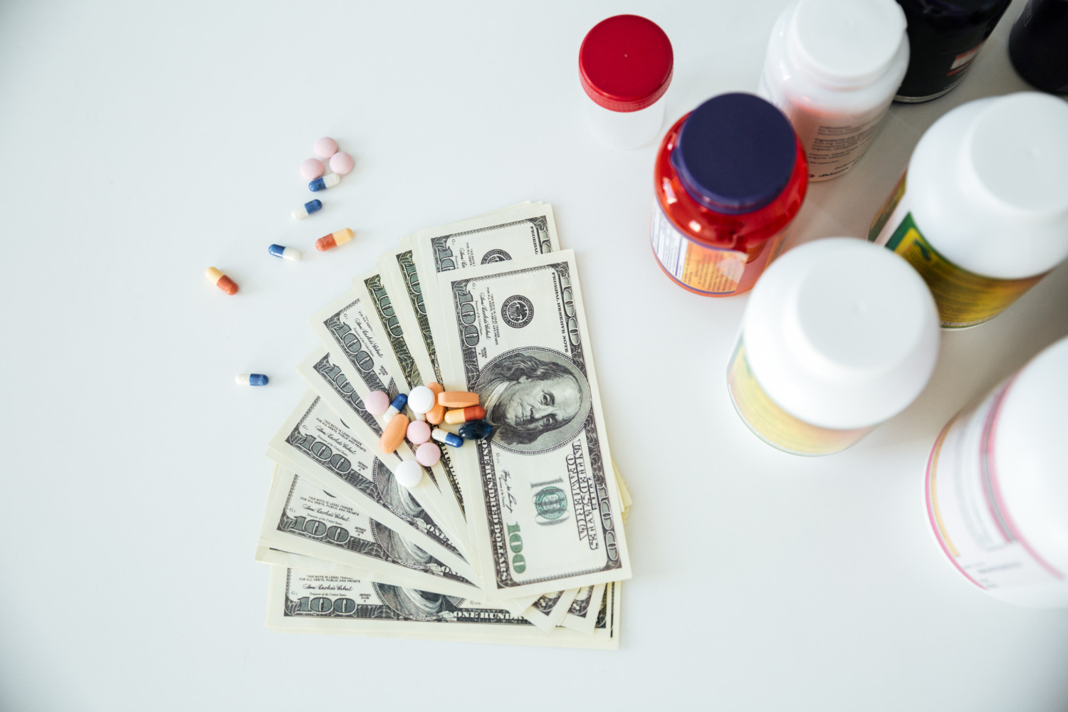 Several prescription pill bottles and pills are on top of U.S. dollar bills on a white surface.
