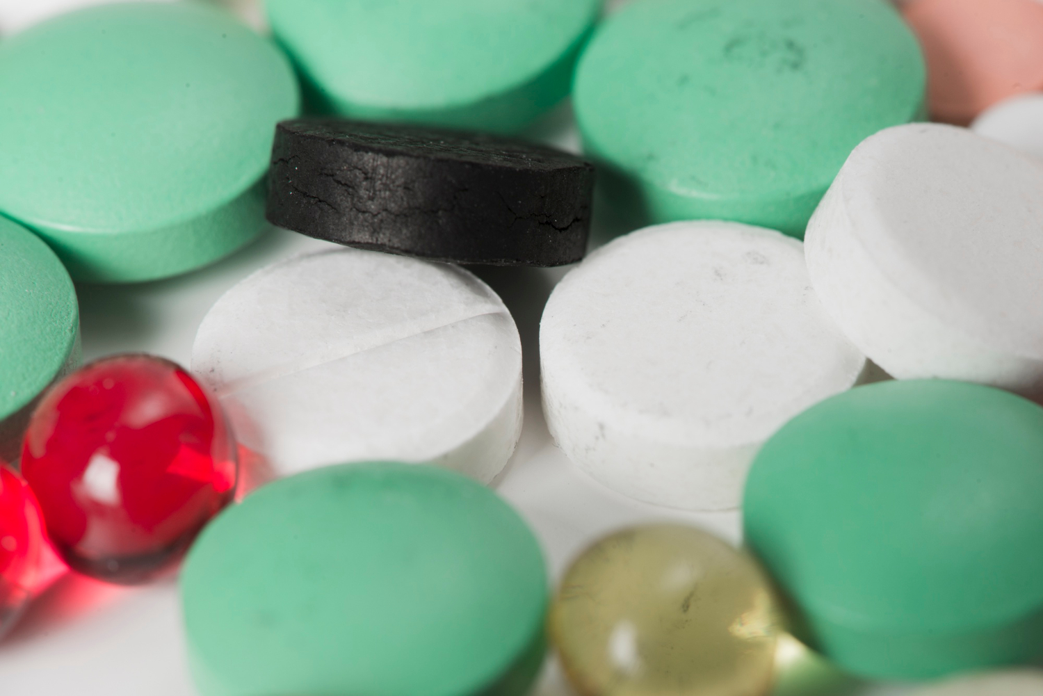 Close-up of assorted pills and tablets in green, white, black, red, and yellow colors on a white surface.