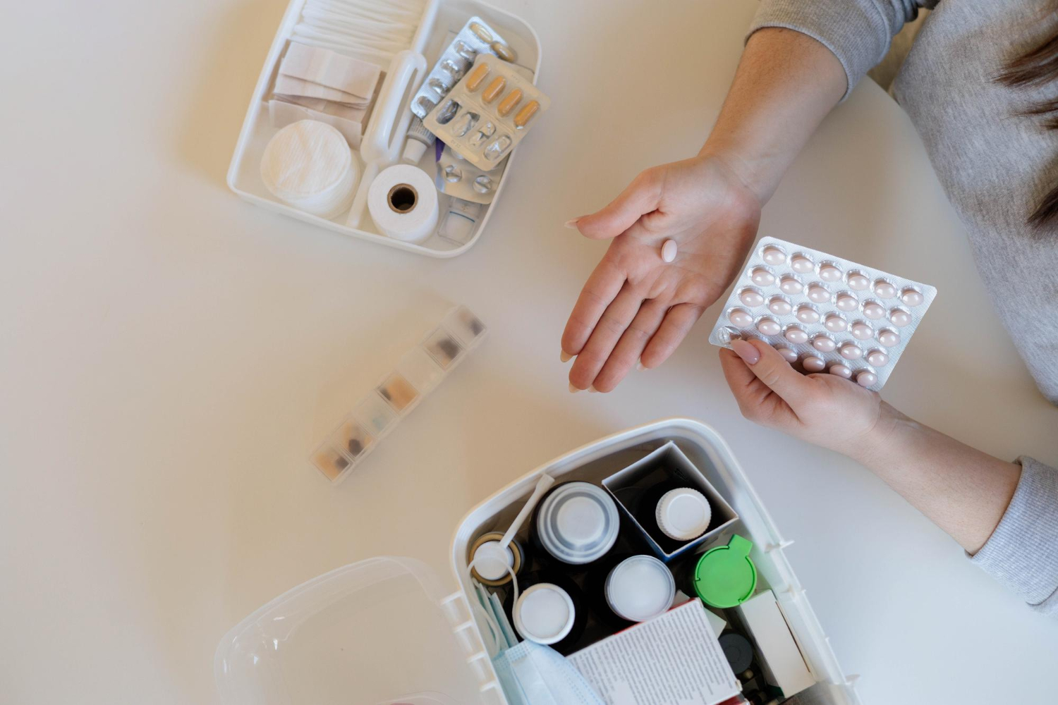 Person holding a pill and blister pack near an open first aid kit with various medical supplies on a table.