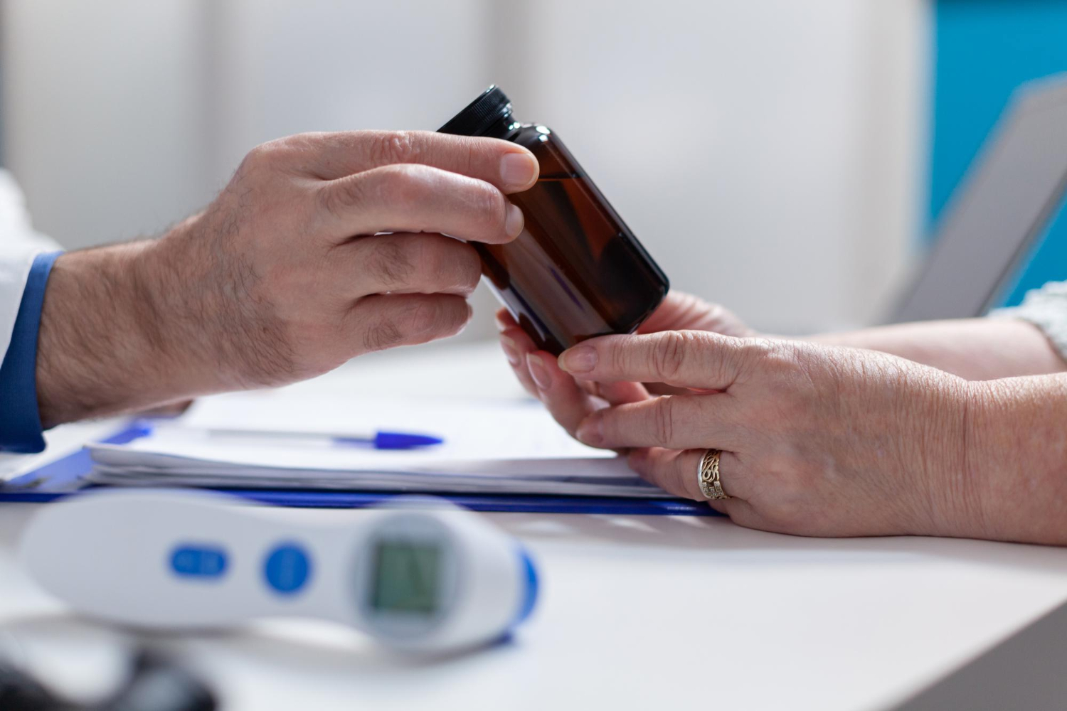 32035 Two people exchange a brown medicine bottle across a desk with medical papers and a thermometer.