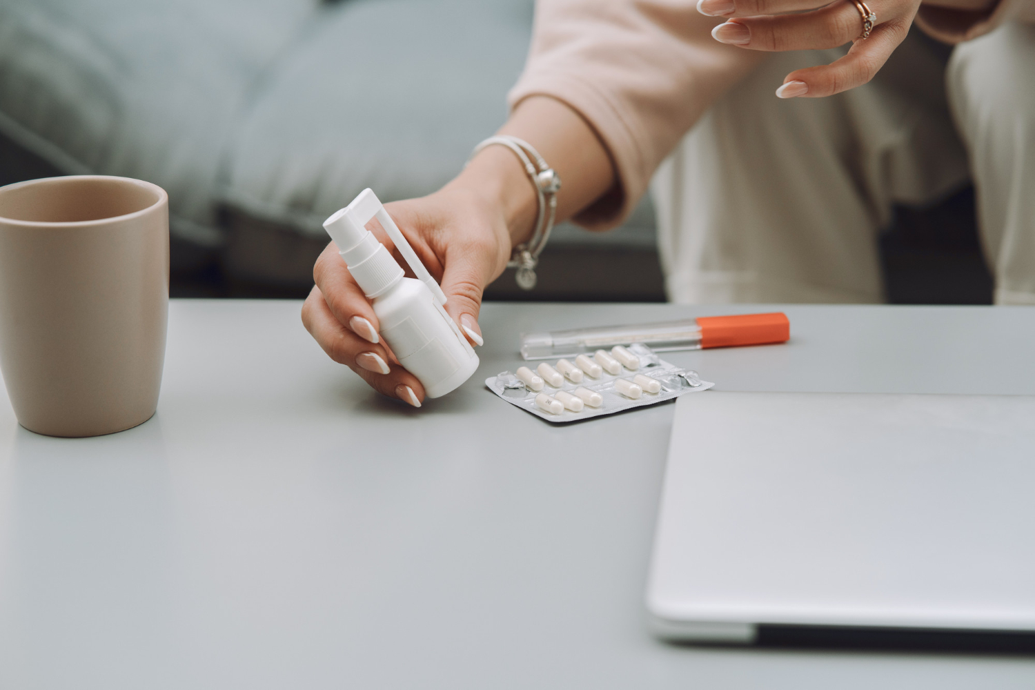 36776 A person holding a pill bottle near a mug, blister pack of pills, pen, and laptop on a table.