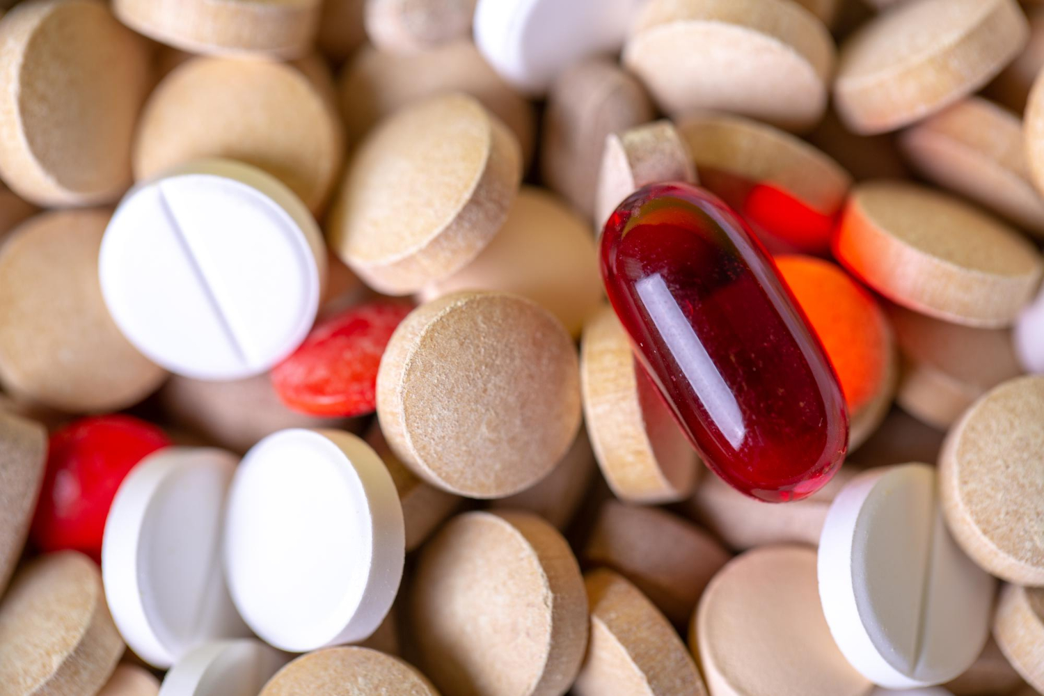 Assorted pills and tablets, including round white, beige, red, and a single oval red gel capsule.