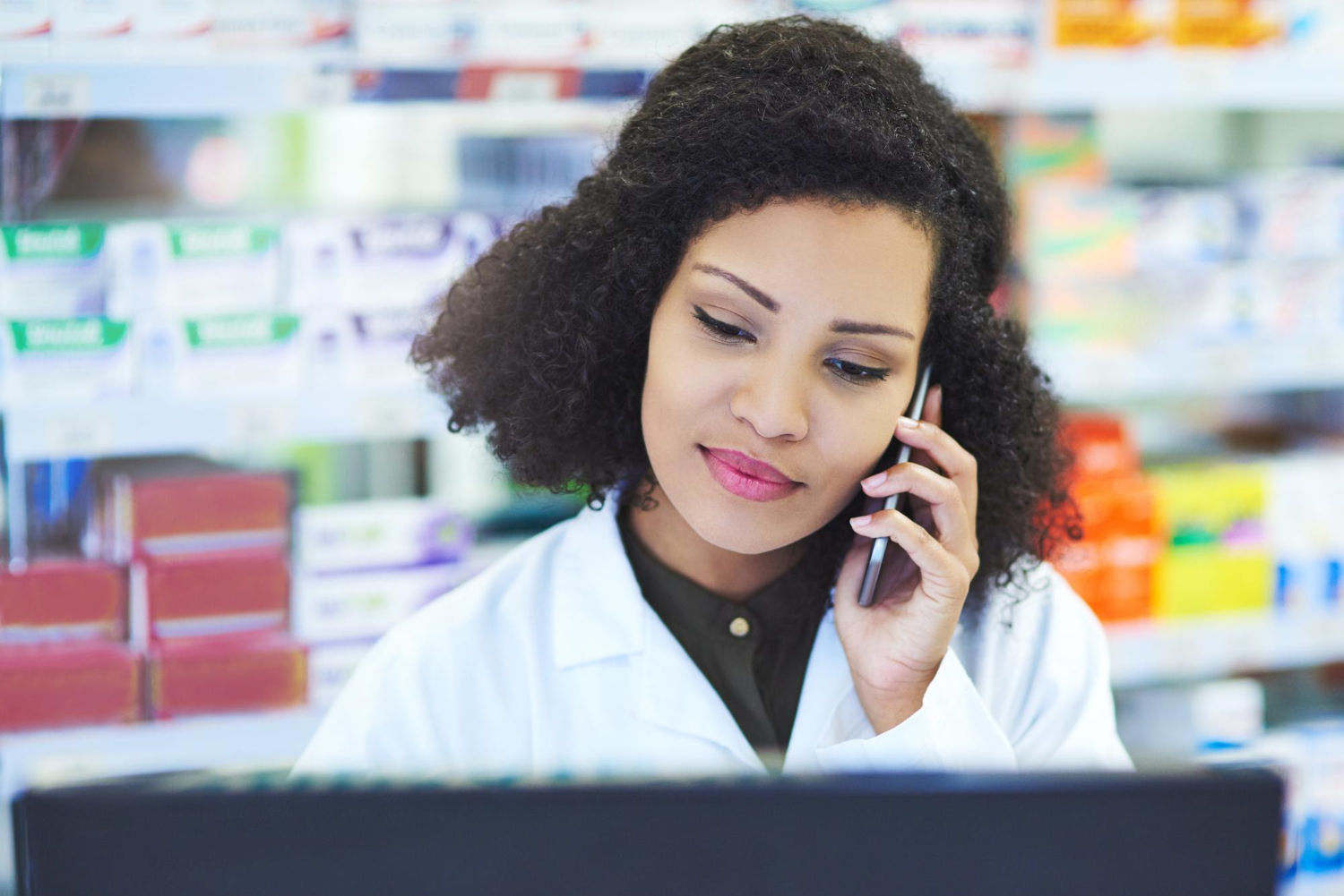 Pharmacist talking on the phone while looking at a computer screen, with shelves of medicine behind her.