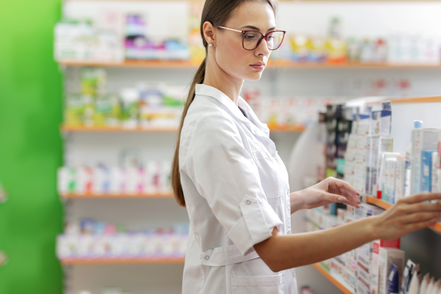 A pharmacist in a white coat organizes medication boxes on shelves in a pharmacy.