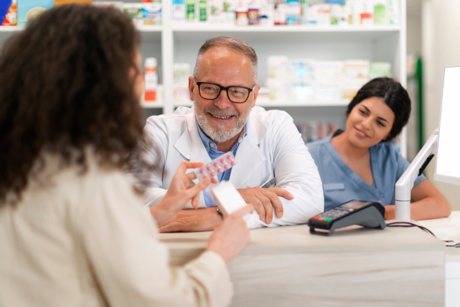 2150600130 A pharmacist and an assistant help a customer holding medication at a pharmacy counter.
