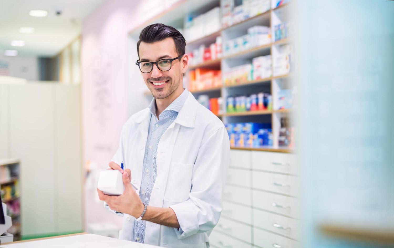 Smiling pharmacist in a white coat holds a medicine box behind the counter in a pharmacy.